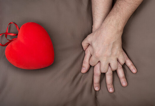 LGBT concept. Men holding hands with passion on the bed near red heart. Hands of couple of homosexual men on brown pillow. Same sex love. Gay relationships. Valentines card for gays