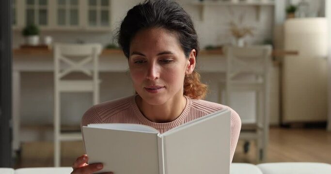 Smiling young beautiful caucasian woman involved in reading favorite bestseller paper book at home, enjoying interesting story, studying indoors, booklover domestic hobby pastime activity concept.
