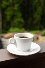 The white mug of dark hot morning coffee on wooden fence with forest background