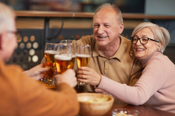 Portrait of smiling senior couple drinking beer in bar and clinking glasses while enjoying night...