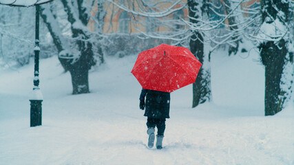 Unrecognizable woman carrying a red umbrella walks along an empty snowy avenue.
