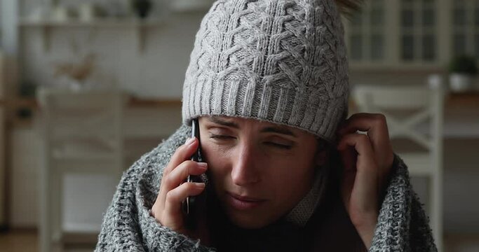 Close up unhappy young woman in warm clothes holding cellphone call, complaining about cold temperature indoors. Stressed lady wearing scarf and cap, feeling first fly symptoms, calling doctor.