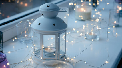 Close-up of burning tea candles in lantern and glasses on sill. Led lights garland. Hygge christmas decoration concept.