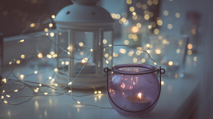 Close-up of burning tea candles in lantern and glasses on sill. Led lights garland. Hygge christmas decoration concept.