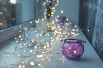 Close-up of burning tea candles in glasses on sill. Led lights garland. Hygge christmas decoration...