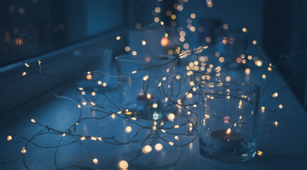 Close-up of burning tea candles in glasses on sill. Led lights garland. Hygge christmas decoration concept.