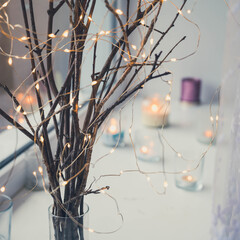 Close-up of led lights garland on branches in glass vase on sill. Burning candles. Hygge christmas decoration concept.