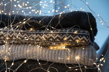 Close-up of led lights garland on stack of cozy knitted sweaters. Hygge warm consept.