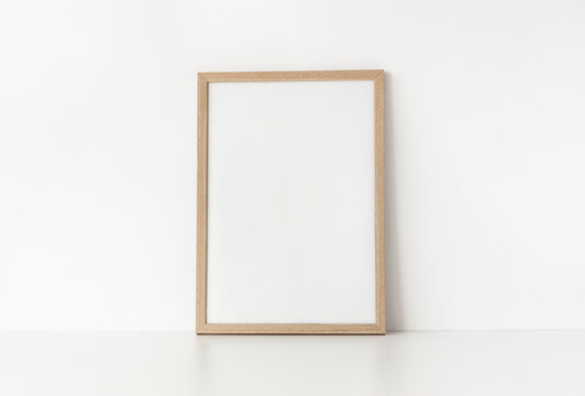 Empty wooden frame stands on white table against wall. Mockup poster frame close up in home interior. 