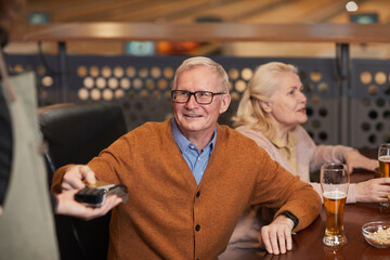 Portrait of smiling senior man paying via NFC in bar while enjoying night out on friends, copy space