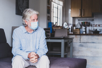 portrait of one old and mature man sitting on the sofa wearing mask to prevent coronavirus indoors - lockdown lifestyle at home.