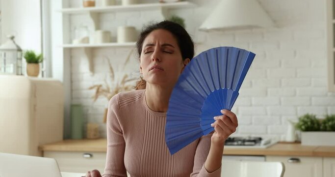 Irritated young caucasian woman using paper fan, blowing fresh air, suffering from hot summer weather or high temperature in kitchen without air conditioner, while working on computer from home.