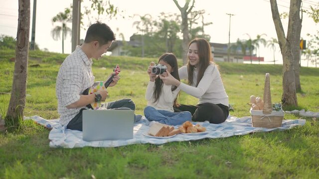 Happy family asian and little girl is playing Ukulele while father take a picture enjoyed ourselves together during picnicking on a picnic cloth in the green garden. Family enjoying sunny fall day.