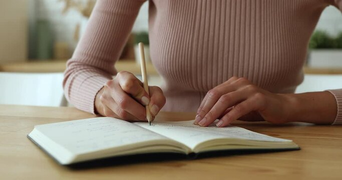 Close up young successful businesswoman planning workday or meetings, writing tasks in paper planner. Focused female student studying, making notes in copybook, sitting at wooden table at home.