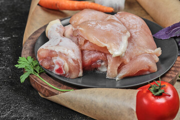 Plate of raw chicken parts with tomato and carrot on dark background