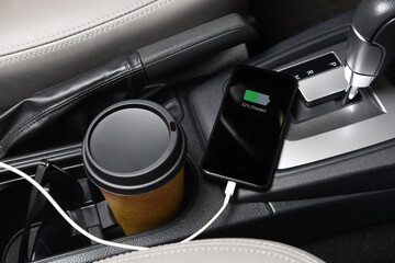 A SmartPhone plugged in the charging port on the console of a modern automobile, closeup with coffee cup and sunglasses.