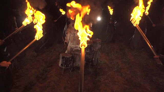 A drummer plays drums in a field at night. Around people with torches.