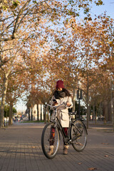 Vertical shot of a young woman on her bicycle using her smartphone. She is wearing winter clothes, and is in a city with autumn weather.
