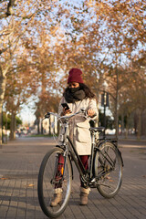 Fototapeta premium Vertical shot of a black woman in winter clothes standing with a bicycle talking on the phone in a city at sunset.