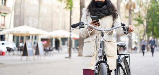 Naklejka premium Panoramic shot of an unrecognizable young African-American woman using a smart phone on her bicycle. She is in a city.