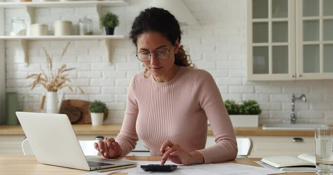 Happy 30s young caucasian woman in eyeglasses calculating paper domestic utility bills, making payment online on computer e-banking applications, feeling satisfied with good financial service at home.