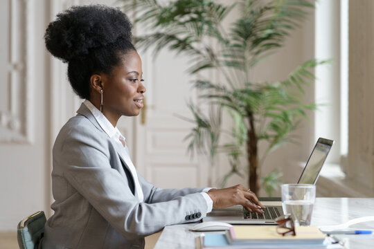 Side view of smiling Afro woman in blazer sitting on desk, remote working at laptop at home office, studying. Confident Black employee female talking in video chat at computer. Distance job.