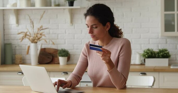 Happy young caucasian woman holding plastic bank card in hands, entering payment information in computer application, purchasing goods or services online, enjoying internet shopping at home.
