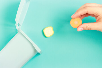 Female hand throws food waste into a plastic trash can on blue background.