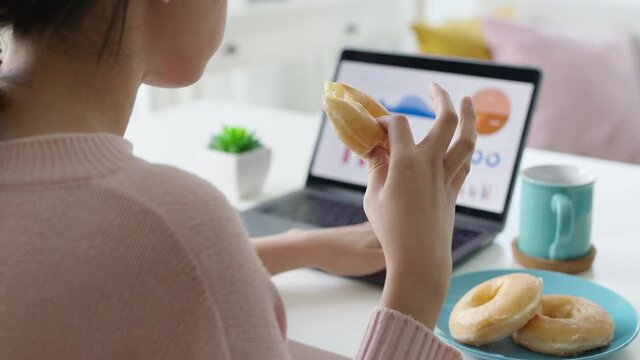 Young attractive beautiful asian female hungry eat doughnut take away snack food with full mouth look at computer notebook at home in busy work from home multitask unhealthy meal lifestyle concept.