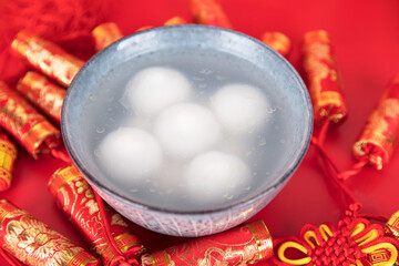 A bowl of Tangyuan or Yuanxiao surrounded by red firecrackers on a red background