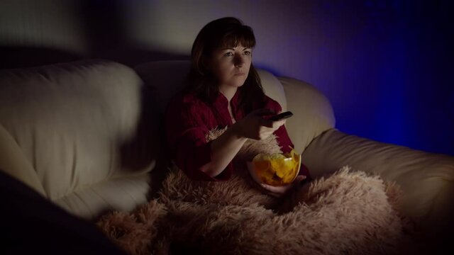 brunette woman sits in sofa with chips at night and watches tv with emotion, switches channels