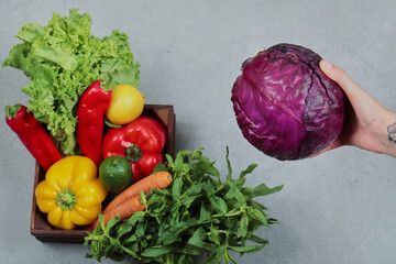 Wooden box of fresh peppers, carrots, lime, lemon and greens and woman holding purple cabbage