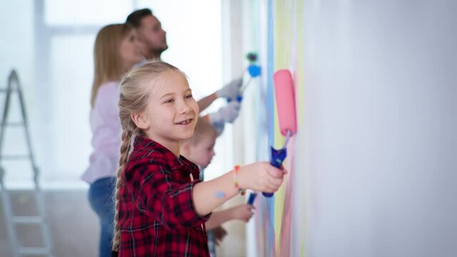 creative kids, cute girl with pink roller in hands paints walls with her brother with down syndrome and parents in background during house renovation