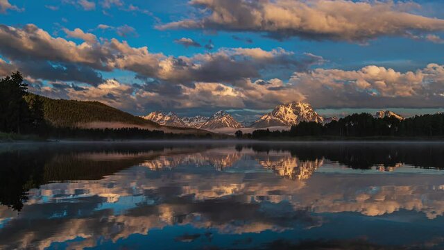 Wide view of beautiful pink and golden sunrise at reflective mirror lake with snow capped mountains and thick clouds flowing by in Grand Teton National Park, Wyoming United States