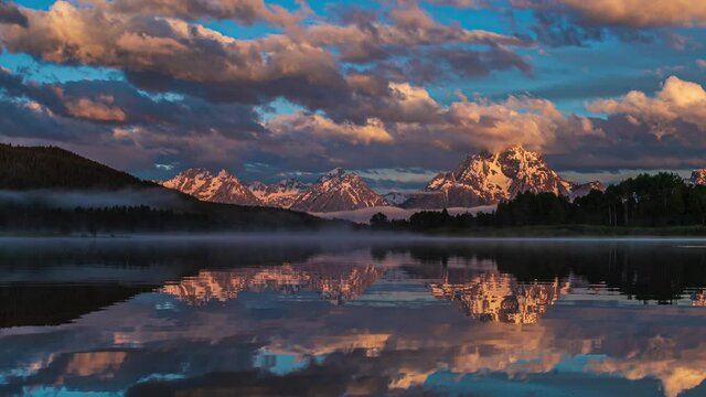 Beautiful pink and golden sunrise at reflective mirror lake with snow capped mountains and thick clouds flowing by in Grand Teton National Park, Wyoming United States