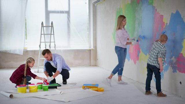 creative family, caring father pours green paint into pallets with daughter while wife with down syndrome son paints walls with construction rollers at construction site indoors