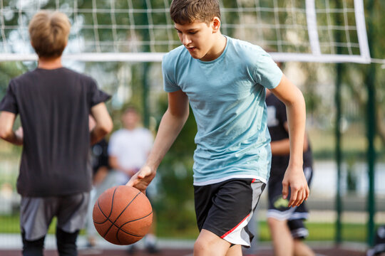 Cute young boy plays basketball on street playground in summer. Teenager in green t shirt with orange basketball ball outside. Hobby, active lifestyle, sport activity for kids.