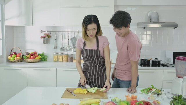 Happy asian couple making health food in kitchen room at home. Young couple make fun and tease while preparing healthy food together indoors. Husband and wife prepare vegetable and fruit for salad.