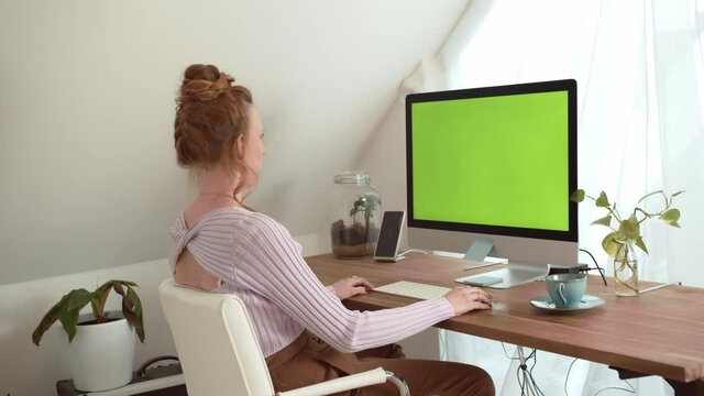 Young beautiful woman sitting in front of a monitor with green screen chroma key. Woman is working at home office in front of a computer.  Advertise Concept for Website, Application or Business. 4K.