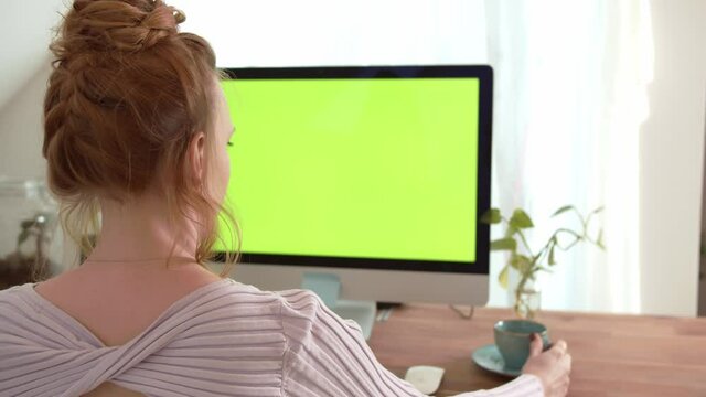 Young women sitting in front of computer with green screen mock-up. Girl drinking Coffee or tea. Over shoulder camera shot. 4k video footage in slow motion.
Concept for online business.