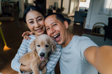 Young adult Asian couple holding a puppy taking a selfie from a phone with home interior in background. 30s mature man and woman with dog pet taking a family photo shots. - Happy group portrait - Powered by Adobe
