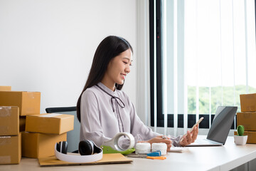 Young Asian woman business owner with many parcel boxes on the table happy online sales job, use your smartphone, get an order from customers, take notes, and make arrangements for delivery by post