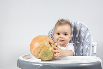 Fototapeta premium A cute little baby holding a fresh pumpkin on a high chair on a white background. Vitamins for children in food.
