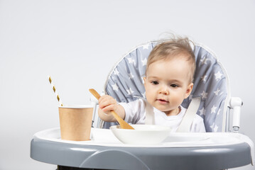 Little beautiful child eating a wooden spoon of yogurt from a plate and a paper cup with a straw with milk on a high gray chair on a white background