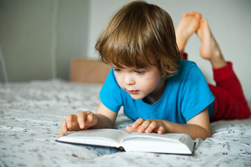 Naklejka premium Little boy lying on the bed and reading a book.