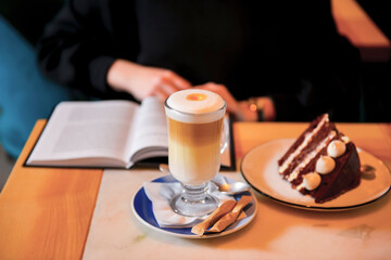 A cup of freshly brewed coffee latte on a table with a book and a piece of chocolate cake. Still life.