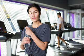 Portrait of sporty woman resting after fitness training in gym standing with bottle of drinking water