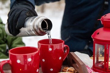 Picnic in winter in the park. The man pours from the thermos into the cup. Two red cups, a candlestick.