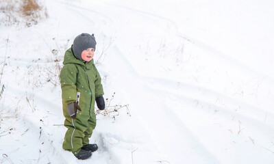 Fototapeta na wymiar A full-length portrait of a child in a green jumpsuit and a grey hat in winter. A boy walks in the snow in winter.