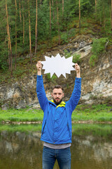 Young man standing on bank of river keeping blank white board in hands. Your text here